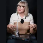 Portrait zeigt TanjaLipp-CVT-Teacher-LAXVOX-Mastertrainer-CopyrightSunlaMahn2024. Auf dem Bild zeigt Tanja Lipp auf eine LAX VOX® Flasche und einen LAX VOX® Schlauch sowie auf das Buch der Complete Vocal Technique