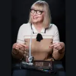 Portrait zeigt TanjaLipp-CVT-Teacher-LAXVOX-Mastertrainer-CopyrightSunlaMahn2024. Auf dem Bild zeigt Tanja Lipp auf eine LAX VOX® Flasche und einen LAX VOX® Schlauch sowie auf das Buch der Complete Vocal Technique