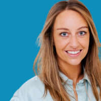 Katrien hat dunkelblond lange Haare, trägt ein blaues Hemd und lächelt in die Kamera.
