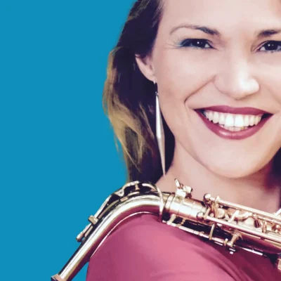 Sabine hat ein Saxophon auf der Schulter und lächelt in die Kamera.